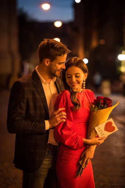 笑容满面的男人拥抱着快乐的女朋友 手里拿着一束玫瑰 卡片上写着爱的题词和心形符号 — 图库照片