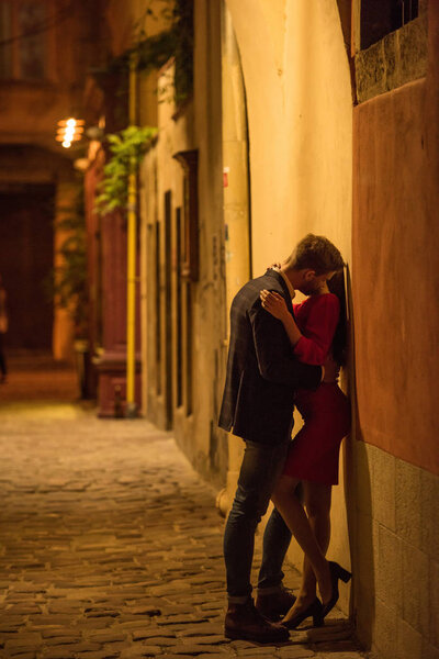 young couple embracing and kissing while standing near wall at night