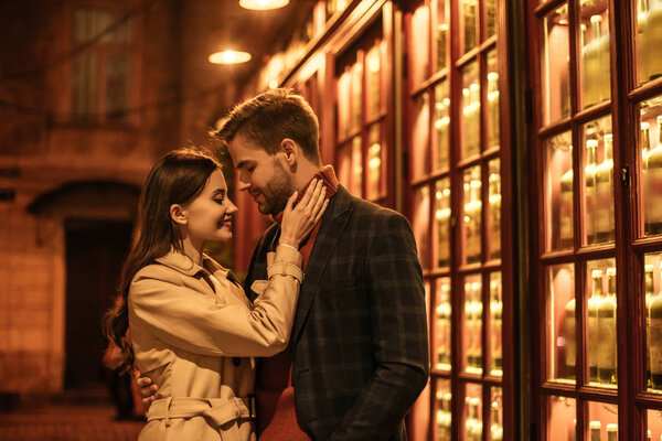 happy girl touching handsome boyfriend whlie standing near shop window at night