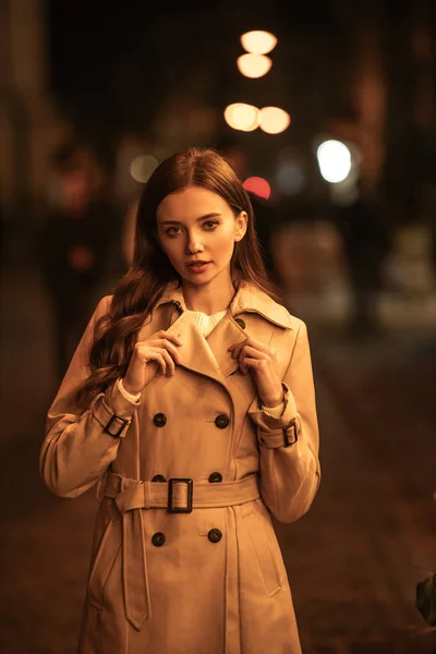 attractive, confident woman in trench coat looking at camera while standing on evening street