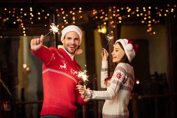 Happy Young Couple Santa Hats Warm Sweaters Holding Sparklers Christmas — Stock Photo, Image