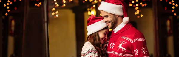 全景拍摄的幸福夫妇穿着温暖的毛衣 头戴圣诞礼帽拥抱在街上 — 图库照片