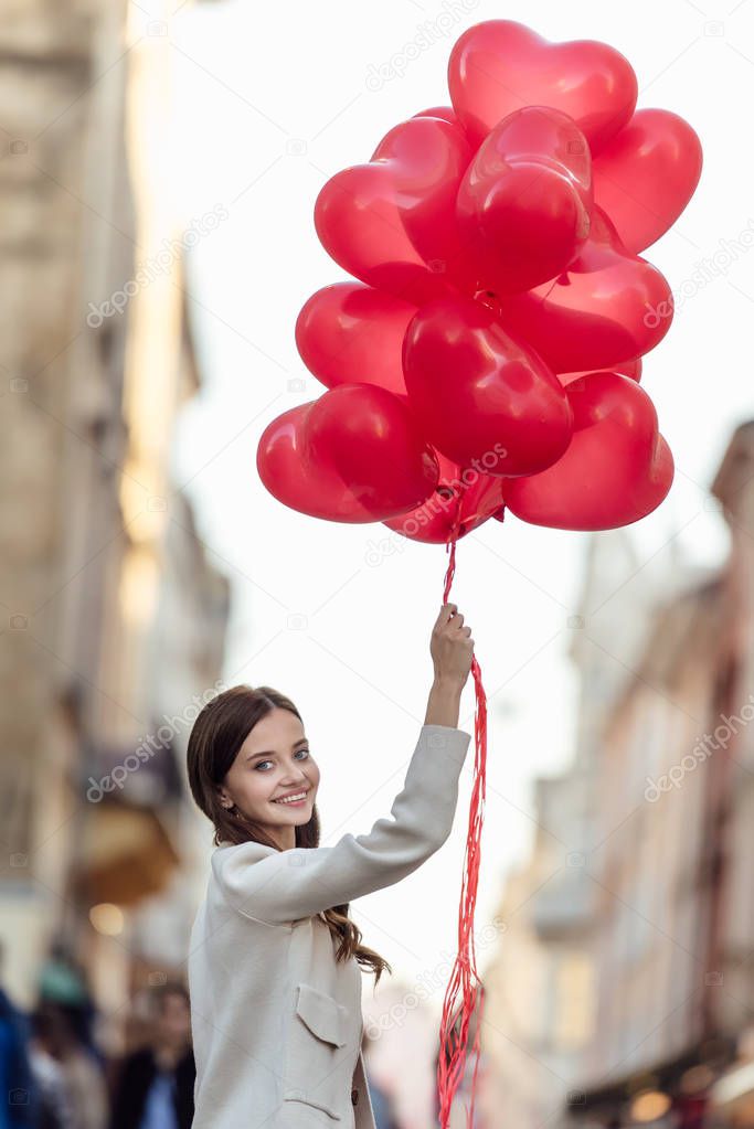 happy girl looking at camera while holding bunch of red heart-shaped balloons on street
