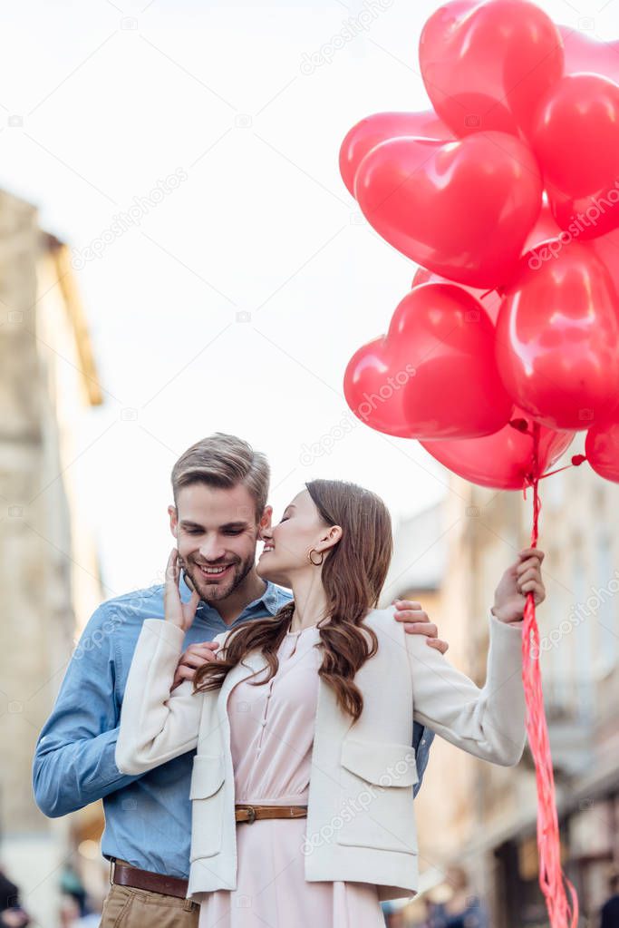 happy girl kissing smiling boyfriend while holding red heart-shaped balloons on street