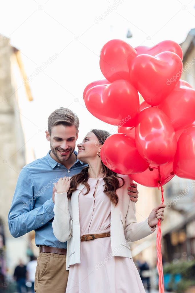 smiling man embracing happy girlfriend holding red heart-shaped balloons on street