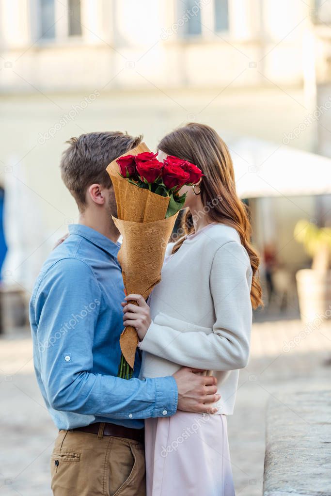 happy couple kissing while hiding behind bouquet of red roses
