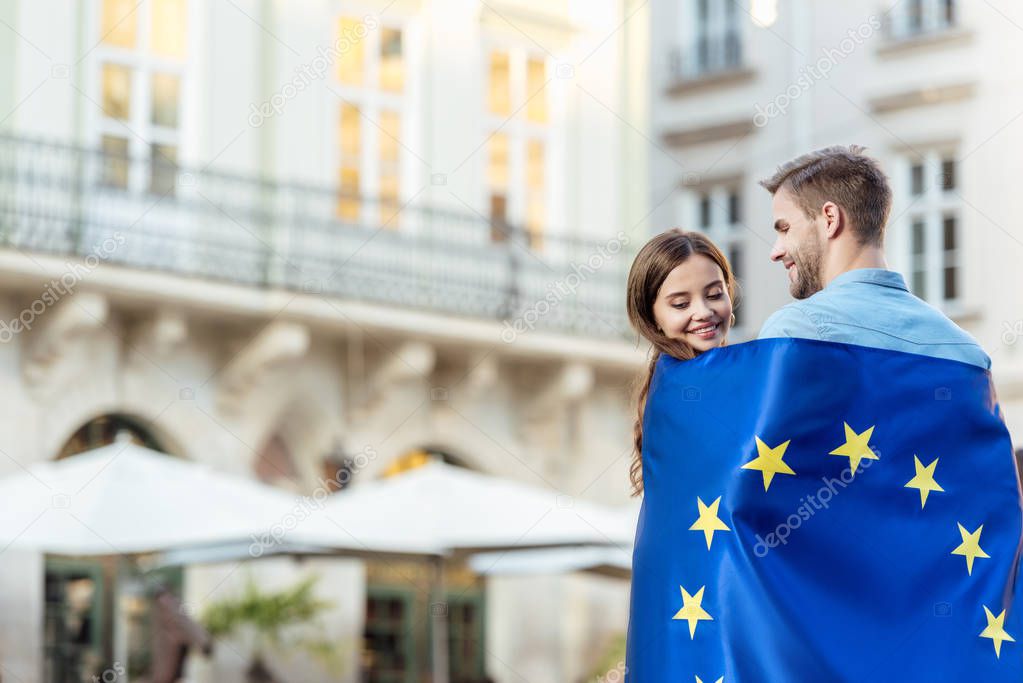 young, smiling couple of tourists wrapping in flag of european union on street