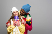 šťastný mezirasový pár v zimním oblečení držení dárek izolované na šedé
