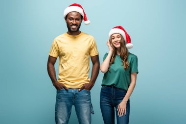 Mavi arka planda Noel Baba şapkalı, ırklar arası gülümseyen çift.