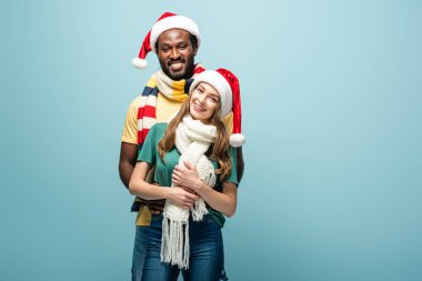 Mutlu ırklar arası çiftler Noel Baba şapkaları ve eşarplarıyla mavi renkte sarılıyorlar.