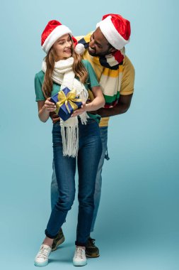 Noel Baba şapkalı ve eşarplı mutlu Afrikalı Amerikalı erkek mavi arka plandaki kız arkadaşına hediye ediyor.