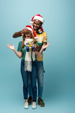Noel Baba şapkalı ve eşarplı mutlu Afro-Amerikan erkeği sürpriz kız arkadaşına hediye ediyor ve mavi arka planda gözleri örtüyor.