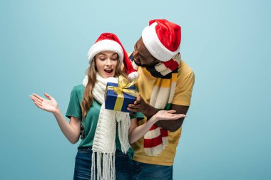 Noel Baba şapkalı ve eşarplı mutlu Afro-Amerikan erkeği mavi elbisedeki sürpriz kız arkadaşına hediye ediyor.