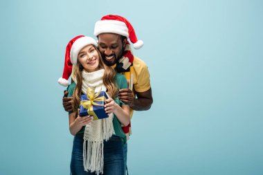 Mutlu ırklar arası çift Noel Baba şapkaları ve eşarplarıyla sarılıyor ve mavi kutuda saklıyorlar.