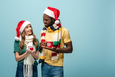 Mutlu ırklar arası çiftler Noel Baba şapkaları ve eşarplarıyla ellerinde mavi renkli kakao bardaklarıyla.