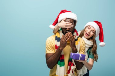 Noel Baba şapkalı ve eşarplı mutlu bir kız Afro-Amerikan erkek arkadaşına bakıyor ve mavi üzerine izole edilmiş hediyeler veriyor.