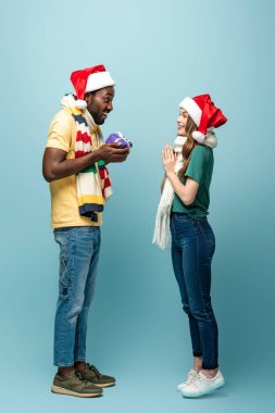 Noel Baba şapkalı ve eşarplı mutlu kız mavi arka planda hediyeli Afrikalı Amerikalı erkek arkadaşına bakıyor.