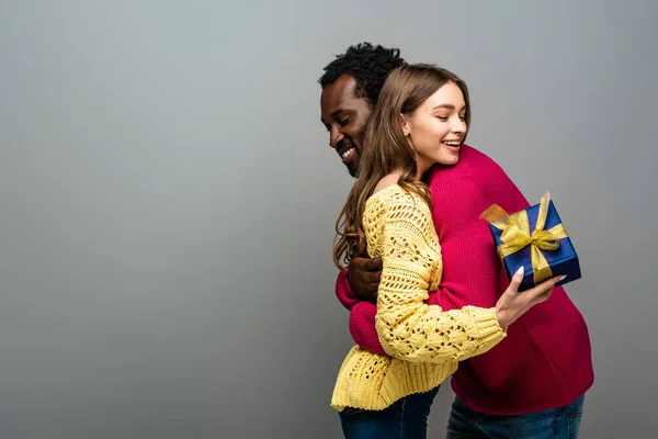 Feliz Casal Interracial Camisolas Segurando Presente Abraçando Fundo Cinza — Fotografia de Stock