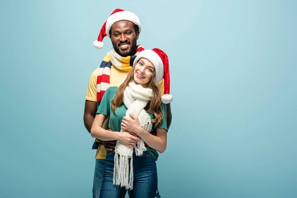 Feliz Interracial Pareja Santa Sombreros Bufandas Abrazo Aislado Azul —  Fotos de Stock