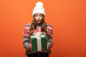 šokované dívka v zimě outfit držení dárek na oranžovém pozadí