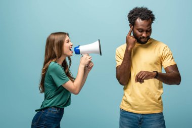angry girl screaming in megaphone at african american boyfriend looking at wristwatch on blue background clipart