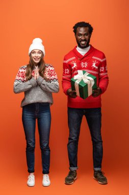 Afro-Amerikan adam elinde Noel hediyesi turuncu arka planda gülümseyen kız arkadaşının yanında duruyor.