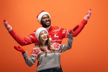 Noel Baba şapkası, eldiven ve Noel süveteri giymiş turuncu arka planda uzanan mutlu bir çift.