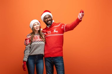 Noel Baba şapkalı, eldivenli ve Noel süveterli mutlu bir çift. Turuncu arka planda el ele sarılıp işaret ediyorlar.