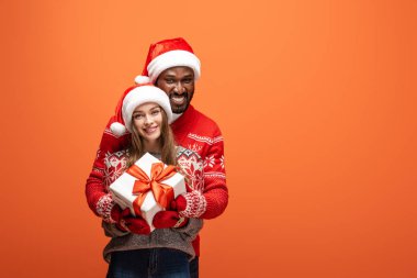 Noel Baba şapkalı ve Noel süveterli mutlu bir çift turuncu arka planda sarılıyor ve hediyelerini tutuyor.