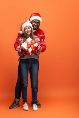 Noel Baba şapkalı ve Noel süveterli mutlu bir çift turuncu arka planda sarılıyor ve hediyelerini tutuyor.