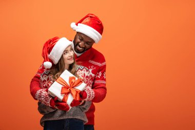 Noel Baba şapkalı ve Noel süveterli mutlu bir çift turuncu arka planda sarılıyor ve hediyelerini tutuyor.