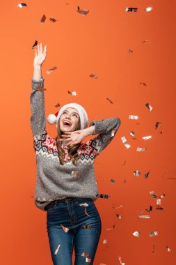 Noel Baba şapkalı mutlu kadın turuncu arka planda konfeti düşüyor.