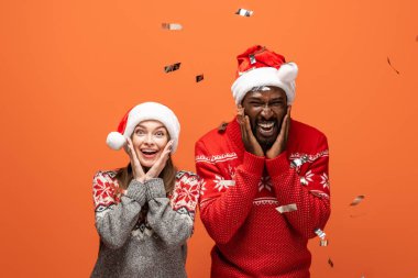 Noel Baba şapkası ve Noel süveteri giymiş turuncu arka planda konfeti altında heyecanlı bir çift.