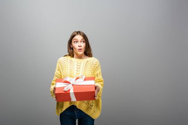 Heyecanlı, mutlu, sarı kazaklı bir kadın gri arka planda hediye kutusu tutuyor.