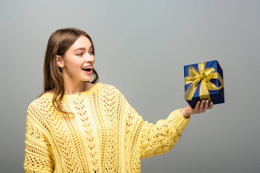 Heyecanlı, mutlu, sarı kazaklı bir kadın gri arka planda hediye kutusu tutuyor.
