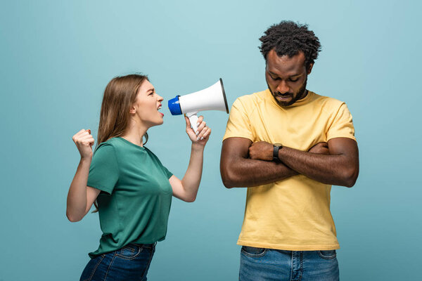 angry girl screaming in megaphone at african american boyfriend with crossed arms on blue background