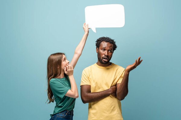 girl holding speech bubble above confused african american man showing shrug gesture on blue background