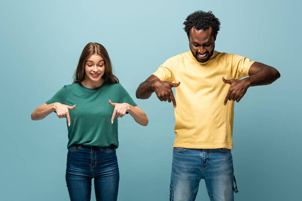 happy interracial couple pointing with fingers down on blue background