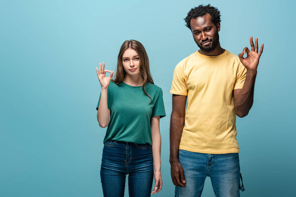 displeased interracial couple showing ok sign on blue background
