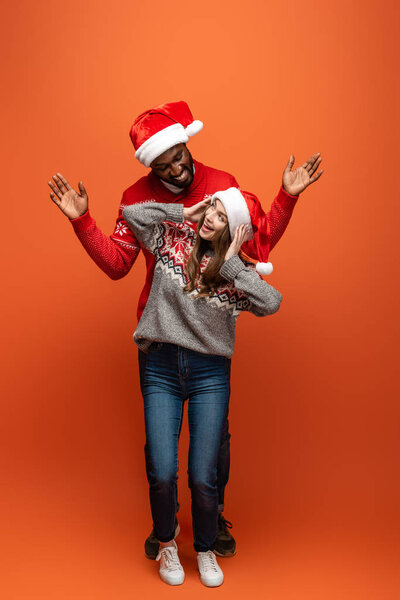 happy interracial couple in santa hats and Christmas sweaters looking at each other  on orange background