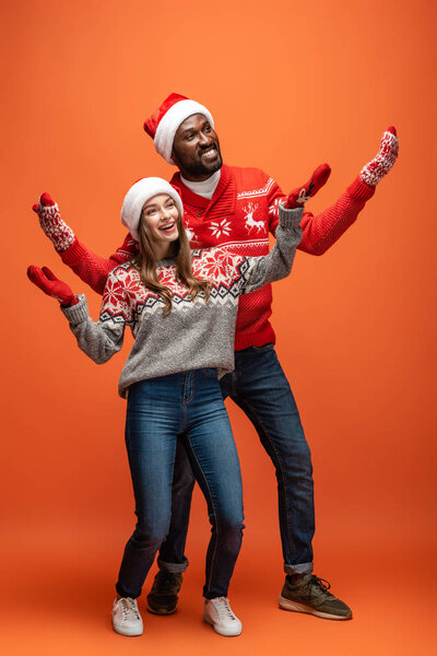 happy interracial couple in santa hats, mittens and Christmas sweaters with outstretched hands on orange background
