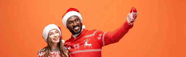 サンタの帽子の幸せな異人種間のカップル ミトンとクリスマスのセーター抱擁し オレンジの背景に手で指して パノラマショット — ストック写真