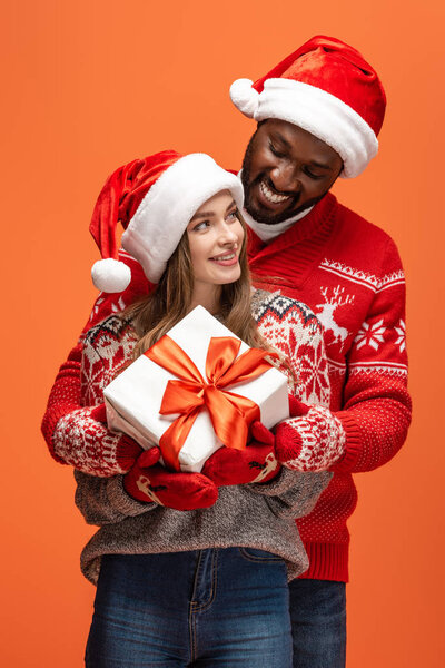 happy interracial couple in santa hats and Christmas sweaters hugging and holding gift on orange background