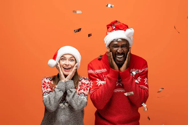 Aufgeregtes Paar Weihnachtsmützen Und Weihnachtspullis Unter Konfetti Auf Orangefarbenem Hintergrund — Stockfoto