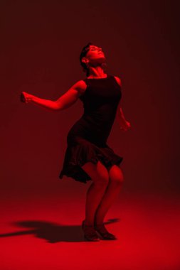Siyah elbiseli şehvetli dansçı kırmızı ışıkla karanlık arka planda tango yapıyor.