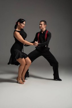 Gri arka planda tango yapan siyah elbiseli iki gülümseyen dansçı.