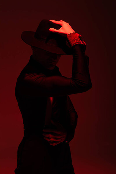 expressive dancer in black clothing and hat performing tango on dark background with red illumination