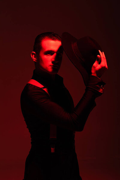 handsome, confident tango dancer looking at camera while holding hat on dark background with red illumination