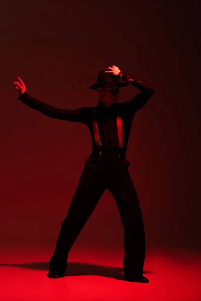 Elegancki Tancerz Dotykający Kapelusza Podczas Wykonywania Tanga Ciemnym Tle Czerwonym — Zdjęcie stockowe