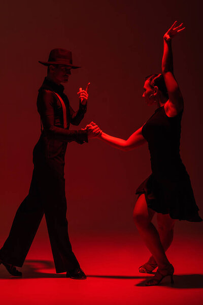 elegant couple of dancers in black clothing performing tango on dark background with red illumination